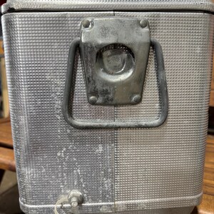 May include: A vintage silver metal cooler with a textured surface and a latch closure. The cooler has a handle and a spigot. The cooler is likely used for keeping drinks and food cold.