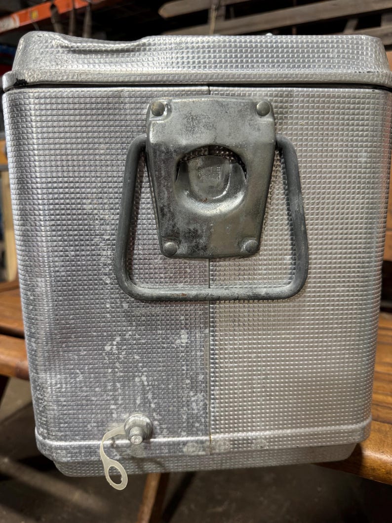 May include: A vintage silver metal cooler with a textured surface and a latch closure. The cooler has a handle and a spigot. The cooler is likely used for keeping drinks and food cold.