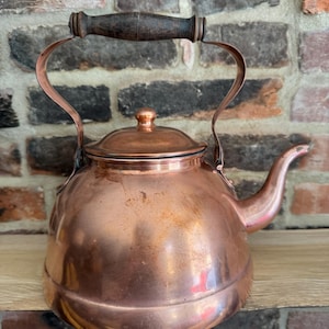 May include: A copper tea kettle with a wooden handle and a curved spout. The kettle has a round body and a lid with a small knob. The copper has a warm, reddish-brown color and shows signs of age.