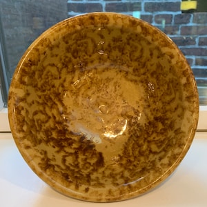 Pode incluir: Uma tigela de cerâmica amarela com um esmalte marrom manchado. A tigela é redonda e tem uma borda ligeiramente alargada.