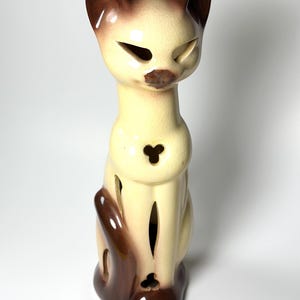 May include: A ceramic cat figurine in a seated position, featuring a cream-colored body with brown accents on the ears, tail, and face. The cat has cut-out eye and body details. The figurine is approximately 10 inches tall.