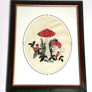 May include: Embroidered artwork featuring red and white spotted mushrooms, green foliage, and red berries. The design is set within an oval frame, framed by a dark green and red wooden frame. A decorative piece for home decor.