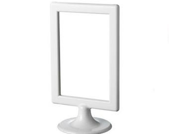 May include: A white, rectangular picture frame with a stand. The frame is empty, with a clean, minimalist design. The stand is round and supports the frame, creating a tabletop display.
