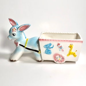 May include: A ceramic donkey planter in light blue and white, pulling a white cart. The cart is decorated with a pink border and illustrations of a blue elephant, a rattle, a yellow giraffe, and a pink flower. The donkey has pink ears and a pink bow.