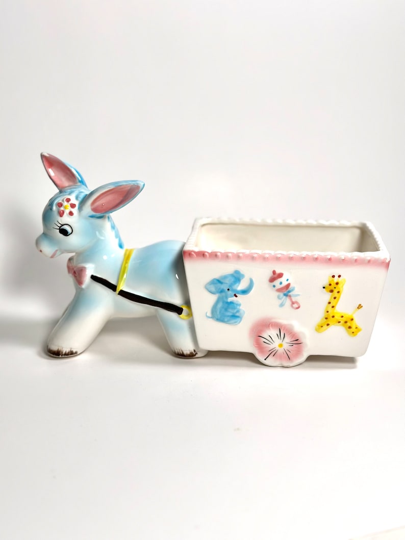 May include: A ceramic donkey planter in light blue and white, pulling a white cart. The cart is decorated with a pink border and illustrations of a blue elephant, a rattle, a yellow giraffe, and a pink flower. The donkey has pink ears and a pink bow.