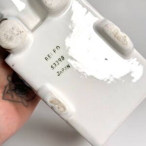 May include: White ceramic rectangular object with four rounded feet. The bottom of the object has the text "REI PO 53398 JAPAN" printed in black.