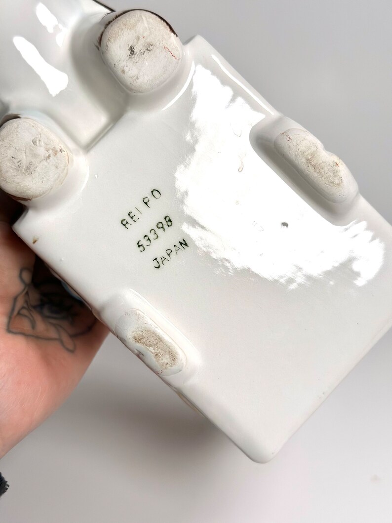 May include: White ceramic rectangular object with four rounded feet. The bottom of the object has the text "REI PO 53398 JAPAN" printed in black.