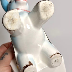 May include: A ceramic donkey figurine, viewed from below, with a white body, blue ears, and a pink bow. The figurine has brown accents on its hooves and is resting on a white rectangular base. The underside of the figurine shows wear.