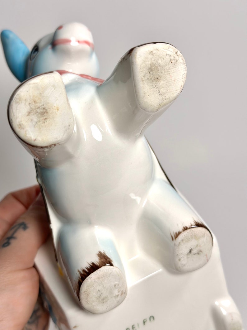 May include: A ceramic donkey figurine, viewed from below, with a white body, blue ears, and a pink bow. The figurine has brown accents on its hooves and is resting on a white rectangular base. The underside of the figurine shows wear.