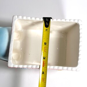 May include: A rectangular, off-white ceramic container with a scalloped edge, measuring approximately 4 inches in length. The container has a light pink and blue trim. A yellow measuring tape is placed inside the container.
