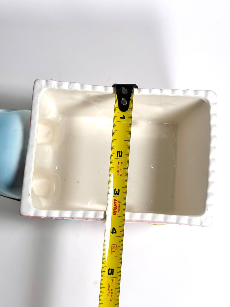 May include: A rectangular, off-white ceramic container with a scalloped edge, measuring approximately 4 inches in length. The container has a light pink and blue trim. A yellow measuring tape is placed inside the container.