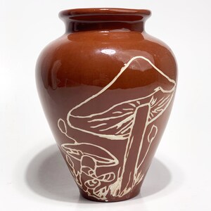 May include: A brown ceramic vase with a glossy finish. The vase features a white outline design of mushrooms. The vase has a rounded body and a slightly flared rim. The design is on the front of the vase.