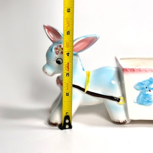May include: A vintage ceramic donkey planter, painted light blue with pink-lined ears and a yellow harness. The planter is approximately 5 inches tall. A white cart is attached, decorated with a blue elephant illustration.