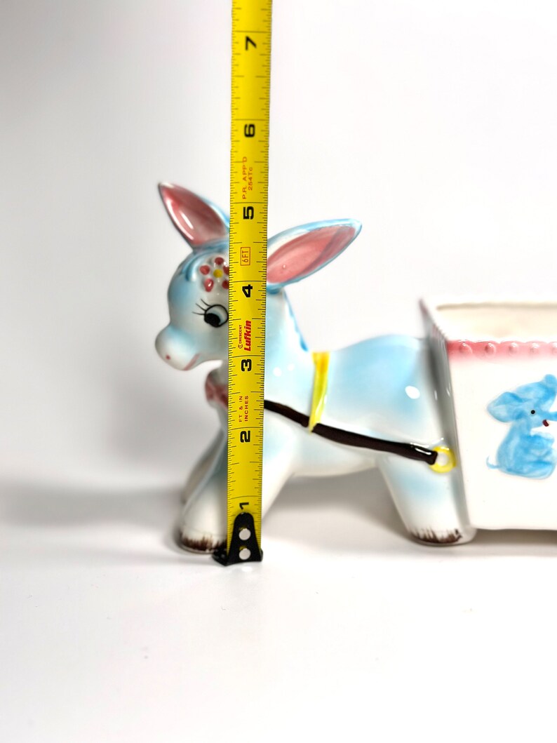 May include: A vintage ceramic donkey planter, painted light blue with pink-lined ears and a yellow harness. The planter is approximately 5 inches tall. A white cart is attached, decorated with a blue elephant illustration.