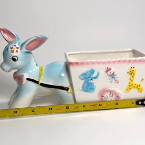 May include: A ceramic donkey planter, painted light blue with pink and yellow accents. The planter has a white rectangular cart decorated with a blue elephant, rattle, and yellow giraffe. The planter measures approximately 9 inches long.