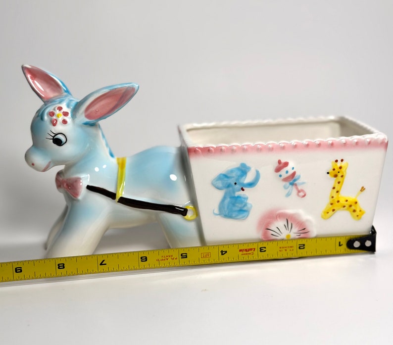 May include: A ceramic donkey planter, painted light blue with pink and yellow accents. The planter has a white rectangular cart decorated with a blue elephant, rattle, and yellow giraffe. The planter measures approximately 9 inches long.