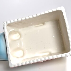 May include: A rectangular ceramic planter with a white interior and a scalloped edge. The exterior features a pink and white floral design. The planter is partially visible, with a light blue section on the left side.