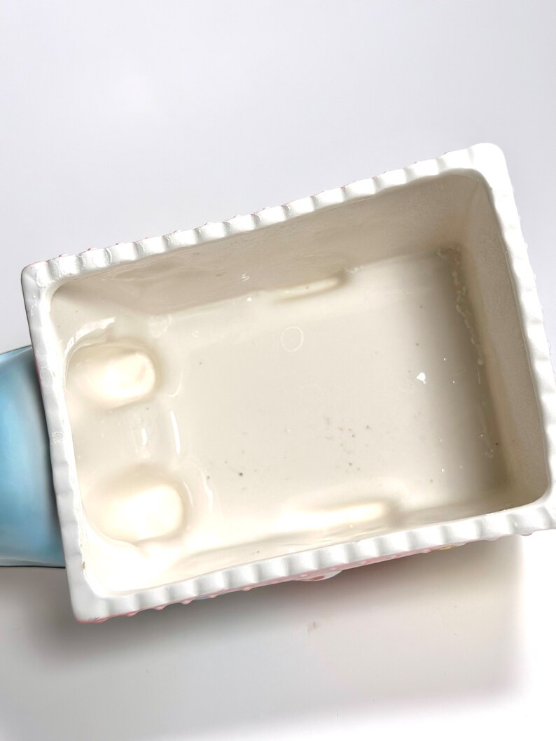May include: A rectangular ceramic planter with a white interior and a scalloped edge. The exterior features a pink and white floral design. The planter is partially visible, with a light blue section on the left side.