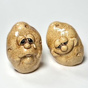 Puede incluir: Dos figuras de cerámica con forma de patata y caras expresivas. Una tiene una lágrima rodando por su mejilla, la otra tiene una expresión gruñona. Ambas son de color canela con ojos oscuros y un acabado brillante.