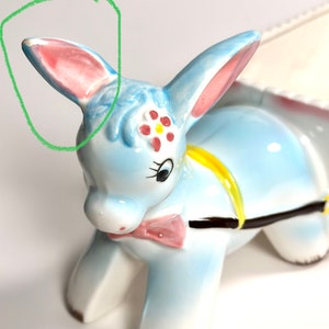 May include: A ceramic donkey figurine in light blue, with pink-lined ears and a pink bow tie. The donkey has a yellow harness and a floral design on its forehead. The figurine is a decorative item.
