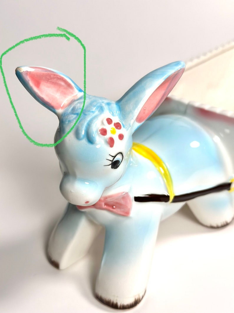 May include: A ceramic donkey figurine in light blue, with pink-lined ears and a pink bow tie. The donkey has a yellow harness and a floral design on its forehead. The figurine is a decorative item.