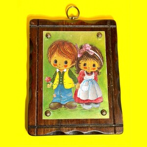 May include: A vintage framed print featuring a cartoon couple holding hands. The boy wears a yellow vest and blue jeans, holding a flower. The girl wears a red dress and white apron. The dark wood frame has a decorative edge and a metal ring for hanging.