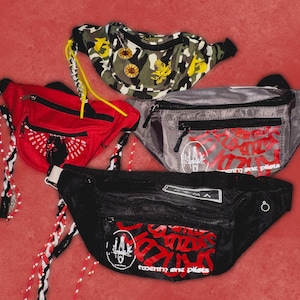 Peut inclure: Quatre sacs banane de différentes couleurs et motifs sur fond rouge. L'un est camouflage, un autre rouge, un gris et un noir. Le sac banane noir porte l'inscription "twenty one pilots" en rouge.
