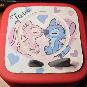 Peut inclure: Un appareil électronique rouge avec un dessus orné d'une image de dessin animé de Stitch et Angel s'embrassant, entourés de cœurs. Le nom "Tara" est écrit en cursive. Deux boutons gris sont visibles.