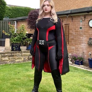 Op de afbeelding: Een vrouw in een zwart en rood superheldenpak met een cape. Ze staat in een grasveld met een bakstenen muur op de achtergrond.