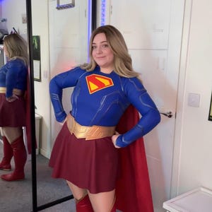 May include: A Supergirl costume featuring a long-sleeved blue top with a red and yellow S-shield emblem on the chest, a dark red skirt, a gold-coloured belt, and a long red cape. The outfit is paired with knee-high red boots.