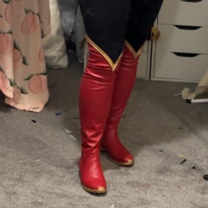 Op de afbeelding: Rode kniehoge laarzen met gouden bies en een gouden zool. De laarzen zijn gemaakt van glanzend materiaal en worden gedragen met zwarte leggings. De laarzen hebben een klassiek ontwerp.