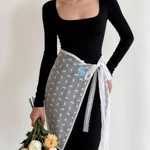 Puede incluir: Un vestido negro de manga larga con cuello cuadrado. Una falda envolvente blanca transparente con un estampado floral y ribete de encaje está atada a la cintura. La envoltura tiene un logotipo azul. Un ramo de flores se sostiene en la mano.