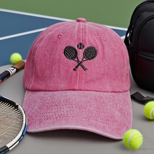 May include: A pink baseball cap featuring an embroidered tennis racket and ball design. The cap has a curved peak and a button on top. Tennis balls and a tennis racket are also visible.