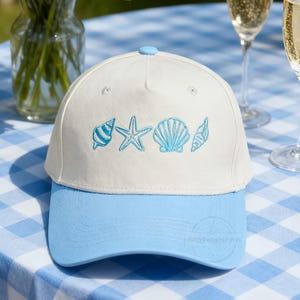 May include: A white baseball cap with a light blue bill and top button. The cap features embroidered light blue seashells and a starfish design. The hat is on a blue and white checkered tablecloth, with champagne flutes and a vase of flowers in the background.