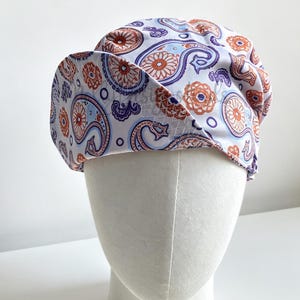 May include: A white scrub cap featuring a repeating paisley pattern in shades of orange, light blue, and dark purple. The hat has a rounded crown and a folded front brim, displaying a detailed print of floral and teardrop-shaped motifs.
