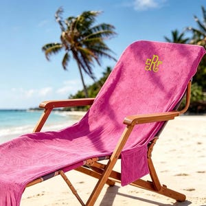 Peut inclure: Une chaise longue en bois recouverte d'une serviette rose vif avec un monogramme jaune brodé. La serviette comporte une poche latérale et recouvre l'assise et le dossier de la chaise, dotée d'un cadre pliant et d'accoudoirs.