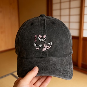 Puede incluir: Gorra de béisbol negra con un diseño bordado con tres caras de fantasmas de dibujos animados en rosa y blanco. El texto japonés "デビル" está encima del diseño. La gorra tiene una visera curva y un aspecto desgastado.