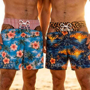 May include: Two pairs of swim shorts featuring white drawstring waistbands. The left pair has a red and white striped waistband above a blue floral print. The right pair has a leopard print waistband above a sunset palm tree print with the name Nathan.