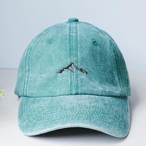 May include: A teal baseball cap with a mountain design embroidered on the front. The cap has a curved brim and a button on top. The hat is made of a soft, textured material.