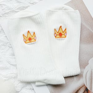 May include: A pair of white socks with a ribbed texture. Each sock features an embroidered crown design in gold, orange, and pink. The socks are displayed on a textured white surface.