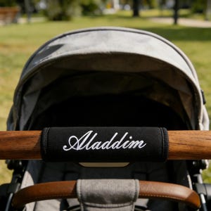May include: A gray stroller with a wooden handle. A black sleeve with the name "Aladdin" embroidered in white is on the handle. The stroller is outdoors with a green grassy background.