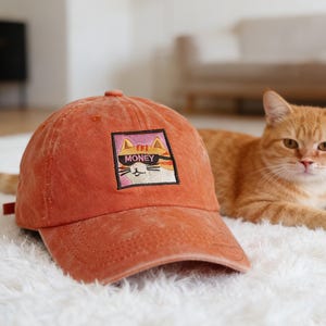 May include: An orange baseball cap with a cat graphic that says "MONEY" on the front. The cat is wearing sunglasses. The cap is sitting on a white surface. An orange cat is lying down in the background.