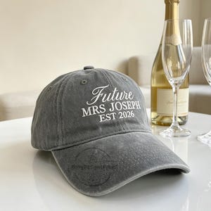 May include: A charcoal grey baseball cap with white embroidered text that reads Future MRS JOSEPH EST 2026. The cap is positioned next to a glass bottle of champagne and two empty clear glass champagne flutes on a smooth white surface.