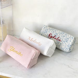 May include: Three personalized cosmetic bags in a row. The first is pink with "Yumiko" embroidered in gold. The second is white with "Melody" in gold. The third is floral with "Ophelia" in red.