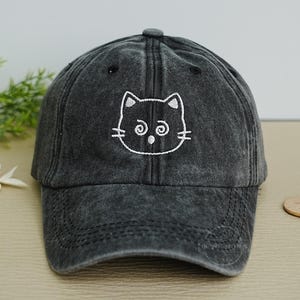 May include: A charcoal gray baseball cap with a distressed finish. The cap features a white embroidered cat face on the front. The cat has large eyes and whiskers. The cap has a curved brim and an adjustable strap.