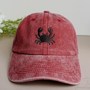 May include: A red baseball cap with a black crab embroidered on the front. The cap has a curved brim and a fabric strap closure. The hat is made of a soft, washed cotton material.
