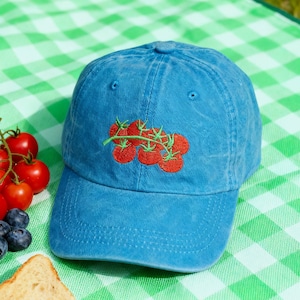 May include: A blue baseball cap with an embroidered design of red tomatoes on a green vine. The hat is sitting on a green and white checkered picnic blanket. Fresh tomatoes, blueberries, and a sandwich are also in the image.