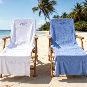 Peut inclure: Deux chaises de plage en bois recouvertes de serviettes personnalisées. La chaise de gauche porte une serviette blanche brodée du prénom Melody en écriture violette. La chaise de droite porte une serviette bleue brodée du prénom Jordan en bleu.