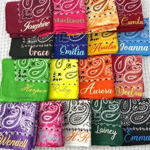 May include: A collection of personalized bandanas in various colors, including red, pink, and blue. Each bandana features a paisley design and a name embroidered in a contrasting color. Names include Josephine, Madison, and Camila.