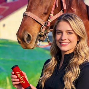 May include: A red and black equestrian crop with the name "Zephyr" in gold lettering. The crop is held in front of a brown horse and a person wearing a black top. The background includes a building with a red roof.